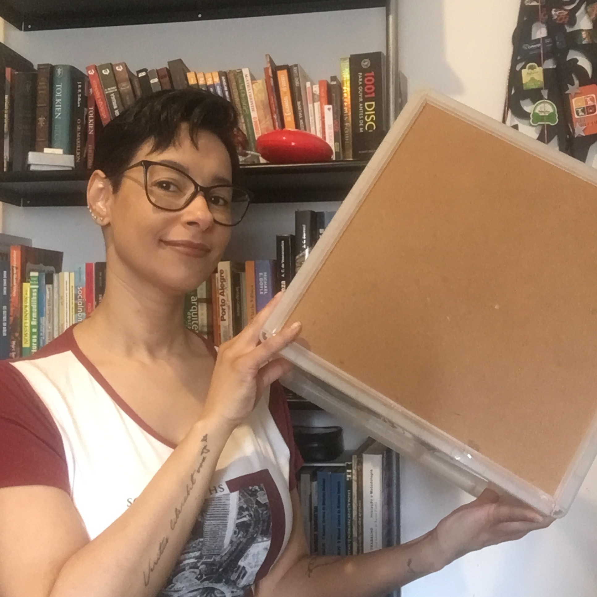 Mulher de óculos segurando uma caixa resistente; estante de livros ao fundo.