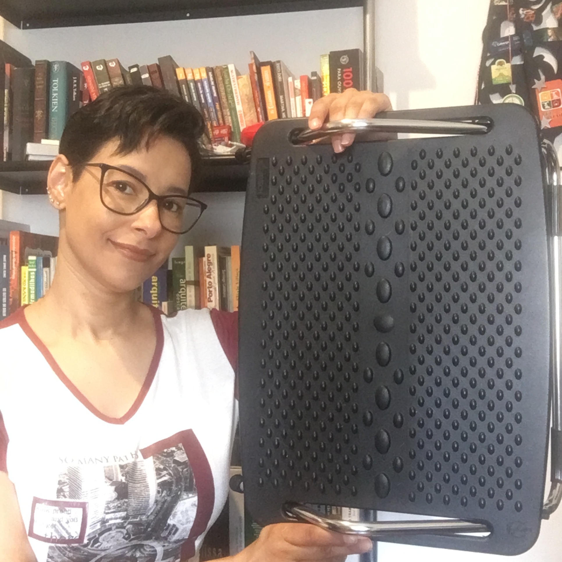 Mulher de óculos segurando um apoio para pés comprado em loja; estante de livros ao fundo.